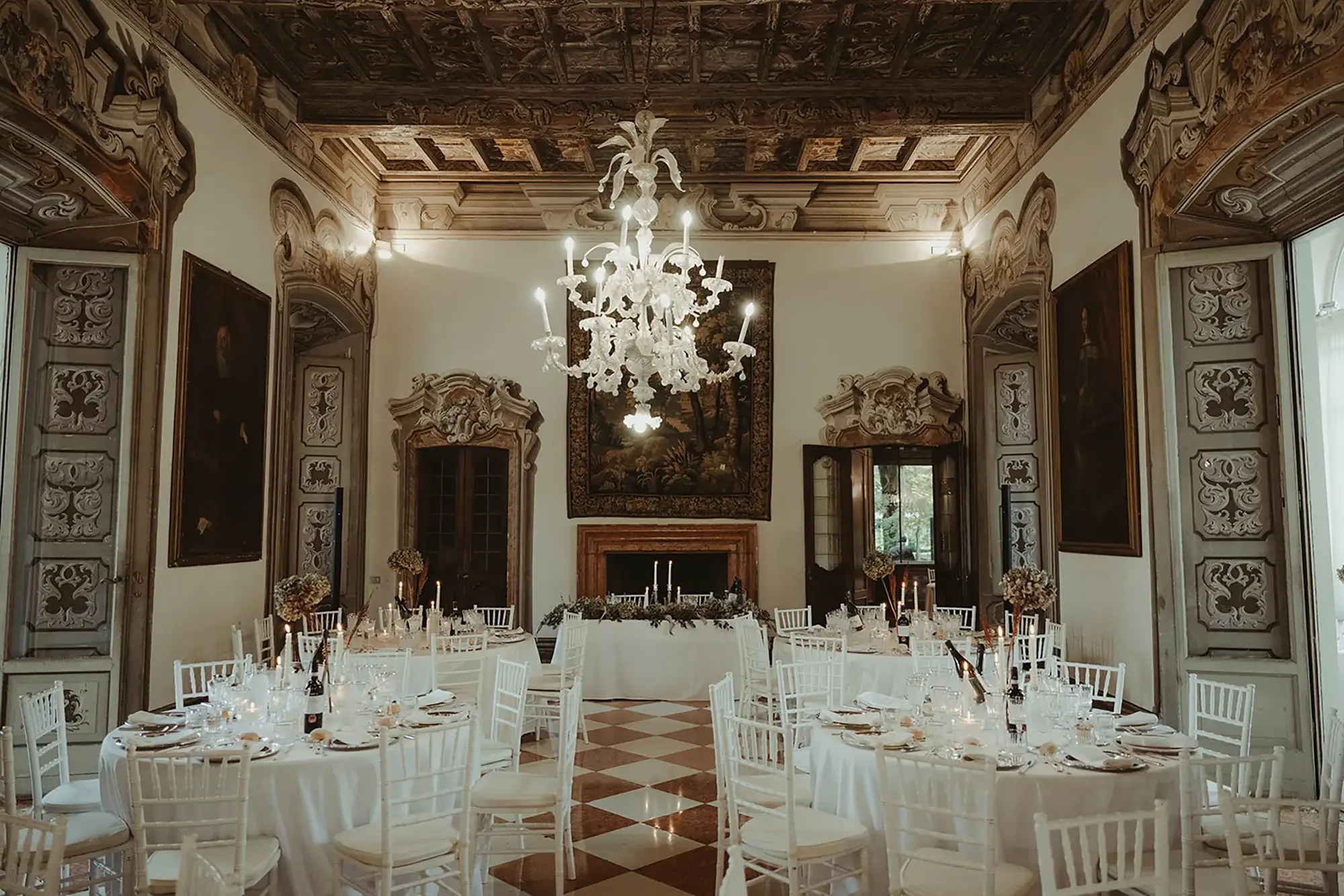 An elegant wedding reception setup in a grand castle room featuring ornate architectural details, including intricately carved door frames, a coffered ceiling, and a central white chandelier. The space is adorned with round tables covered in white tablecloths, surrounded by white chairs, and set with fine dining elements like glassware and candles. A tapestry hangs on the back wall above a decorated table, adding to the luxurious ambiance of the venue.